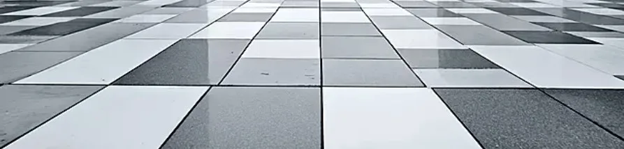A modern floor tiled with rectangular slabs in various shades of white, light gray, and dark gray, arranged in a geometric, checkerboard-like pattern. Some of the tiles appear glossy and reflective, while others have a more matte, textured finish. The image is taken from a low angle, creating a strong sense of perspective as the pattern recedes into the distance.