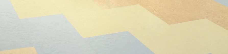 A close-up view of a textured surface, likely flooring or wallpaper, displaying a large, repeating zig-zag (chevron) pattern. The pattern is created using three distinct, muted colors: a pale blue-grey, a soft light yellow, and a warm light tan. The lines of the pattern run diagonally across the frame, and the surface itself has a subtle, granular texture, not perfectly smooth.