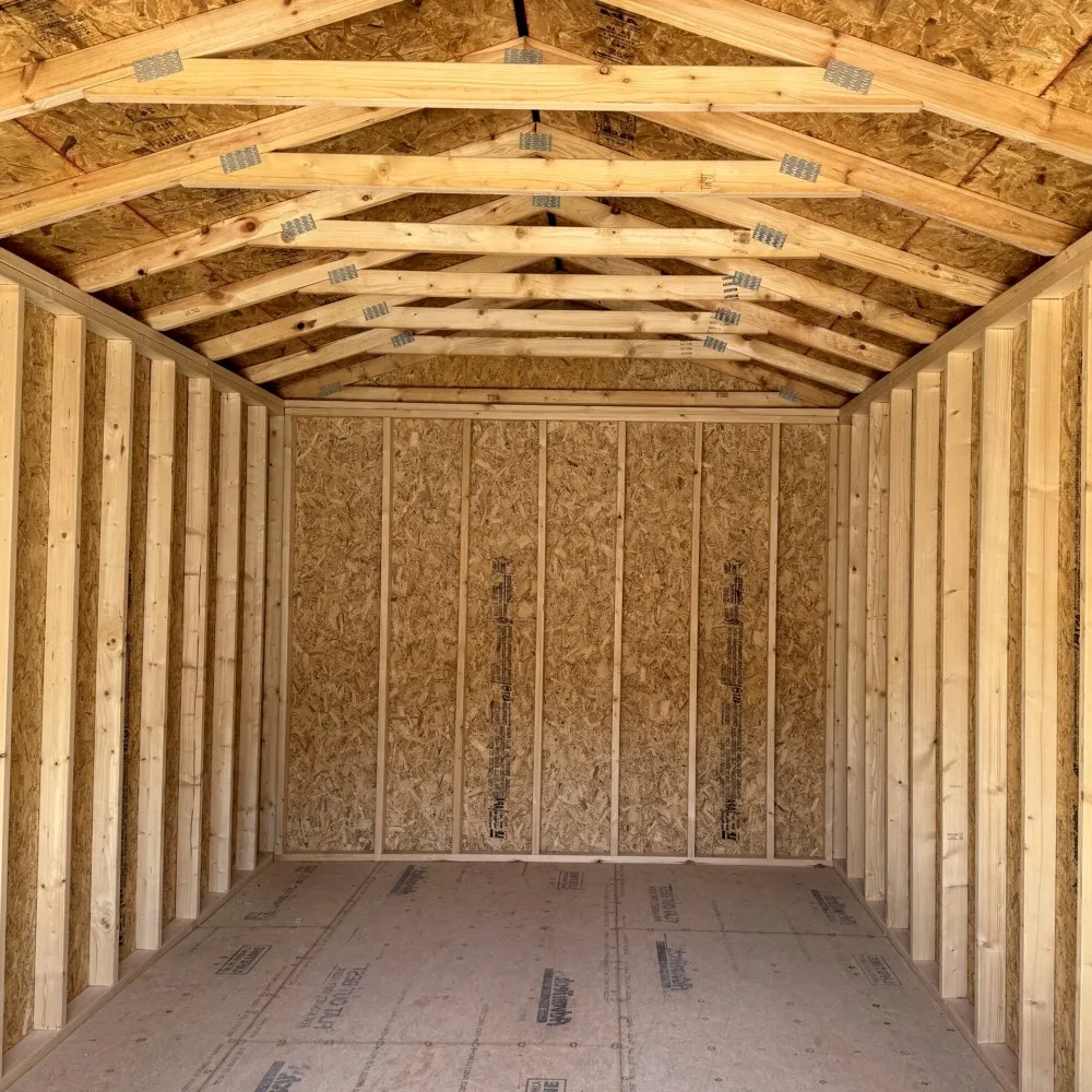 Interior view of an empty, newly constructed wooden building or shed, showcasing its unfinished framework. The gabled roof features exposed wooden rafters and horizontal beams, connected by metal plates, with the underside of OSB sheathing visible. The side walls show vertical wooden studs, while the far wall is clad with vertical panels of Oriented Strand Board (OSB). The floor is made of large sheets of plywood or OSB, with some manufacturer markings visible.