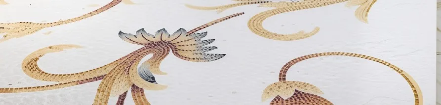 An ornate decorative pattern on a subtly textured white background. It features elaborate curving stems and stylized acanthus-like foliage in shades of golden-brown and terracotta, detailed with fine dotted textures. A prominent central floral motif includes elements transitioning to a muted blue-grey. The overall classical and elegant design suggests a close-up of wallpaper or textile.