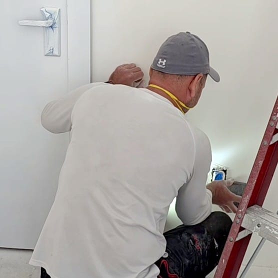 Back view of a person wearing a grey baseball cap and a white long-sleeve shirt, actively sanding or prepping a white interior wall. Their hands are focused on different areas of the wall, one near an electrical outlet with what appears to be a sanding block. A yellow strap, possibly for a respirator or safety glasses, is around their neck. A red step ladder is visible to their right, and a white door frame is on the left.