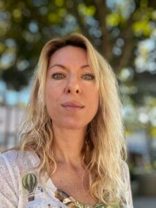 A blonde woman with green eyes gazes directly at the viewer. She has long, wavy blonde hair and is wearing a light-colored top with a green and yellow hot air balloon design. The background is a soft, out-of-focus blur of green foliage and bright highlights, suggesting an outdoor, sunny setting.