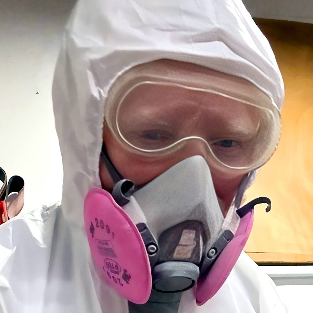 A person wearing a white hooded protective suit, clear safety goggles, and a grey respirator mask with prominent pink filters, looks directly forward.